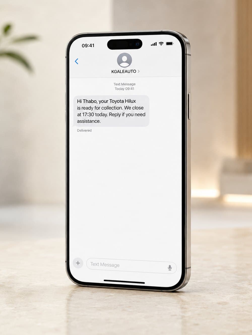 An iPhone on a counter showing a text message from KGALEAUTO reading: Hi Thabo, your Toyota Hilux is ready for collection. We close at 17:30 today.